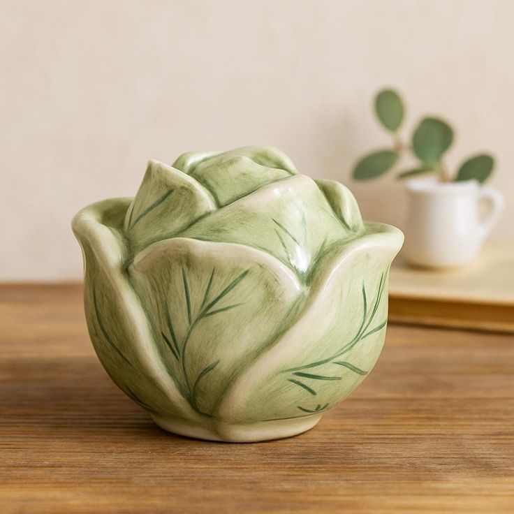 A medium light green ceramic cabbage bowl with a lid sitting on a warm wood shelf with a blurred neutral background and a small plant.