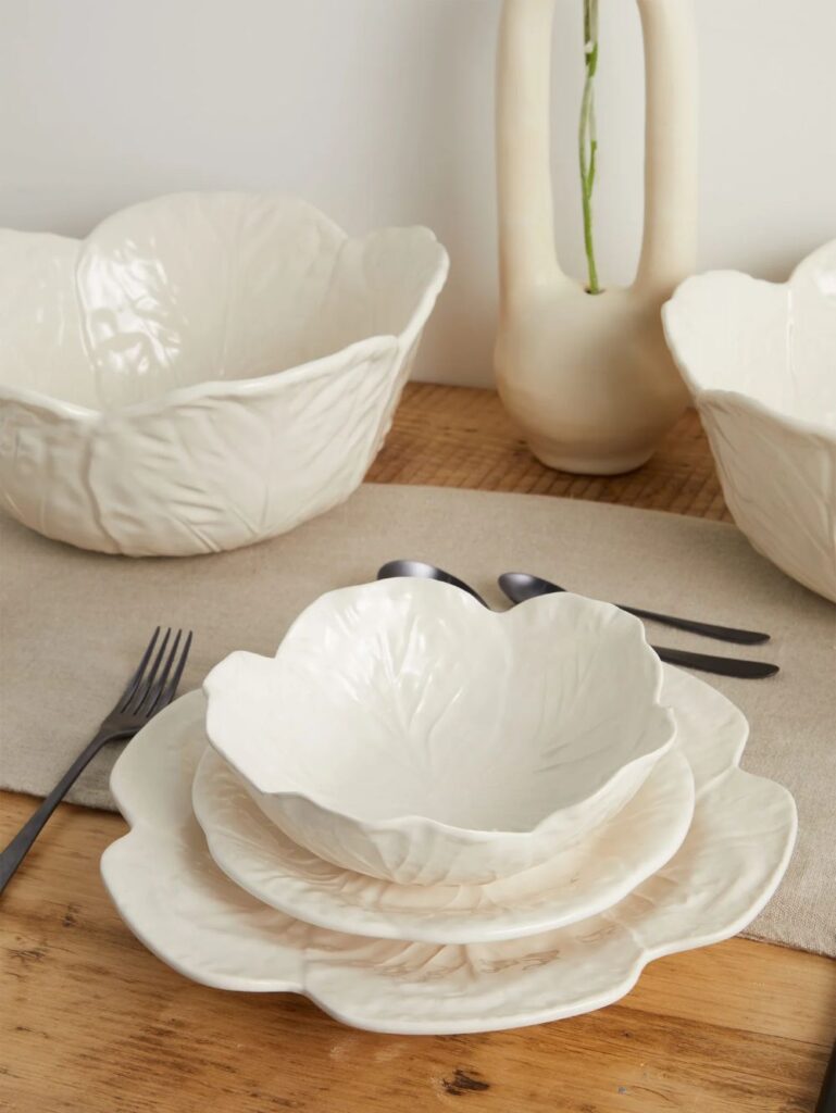 A neutral tablescape featuring a matte white cabbage bowl stacked on matching leafy plates next to a minimalist ceramic vase and black cutlery
